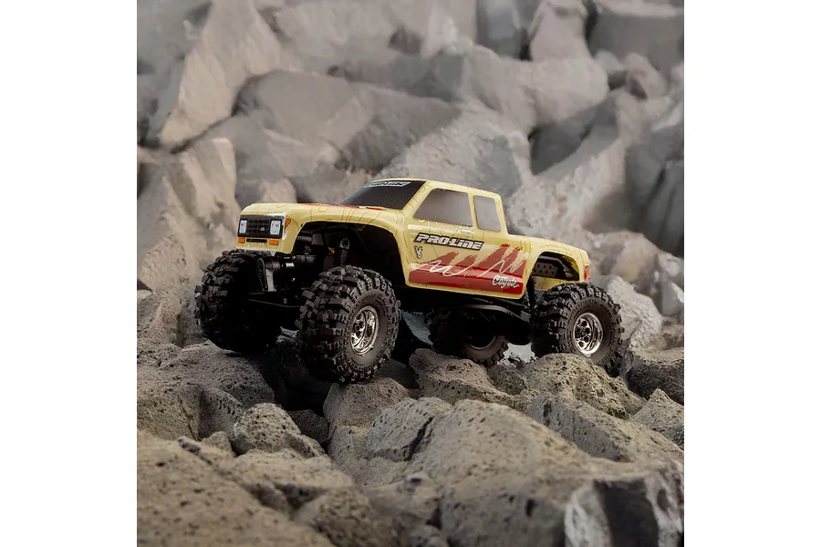 A tan Pro-Line Coyote RC truck body shell with topographical patterns, mounted on a crawler chassis, sitting on a rocky terrain.