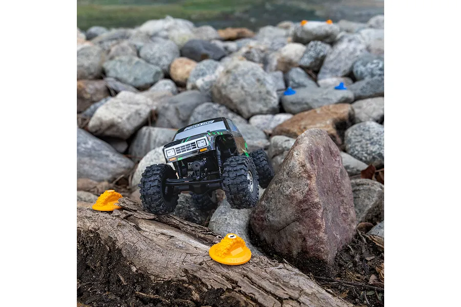 A small RC crawler vehicle driving over a log and rocks, with orange Pro-Line gate markers placed on the terrain.