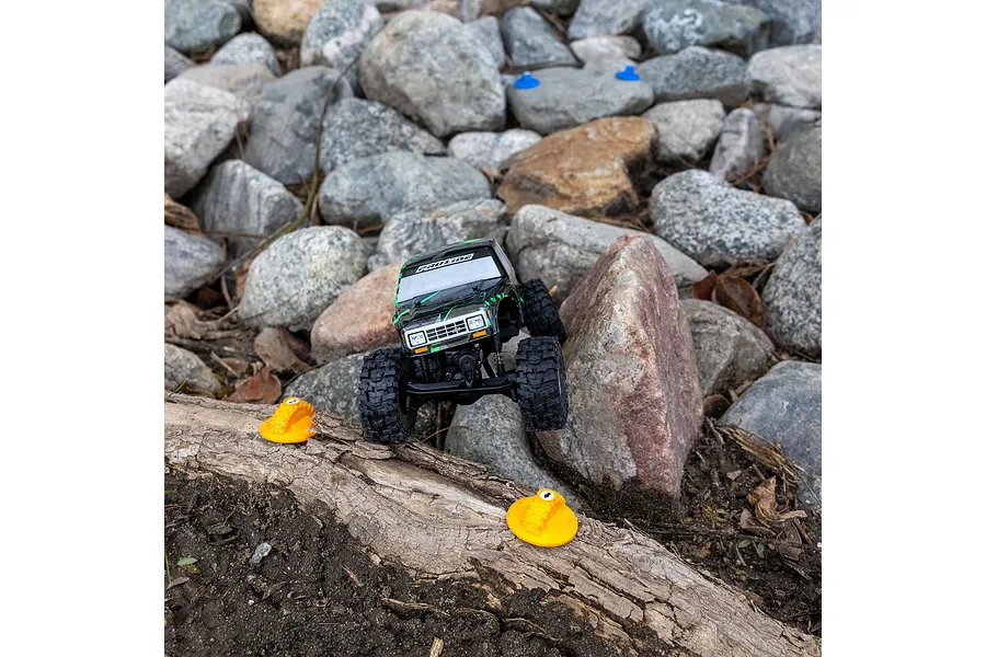 A small RC crawler driving over a log with orange Pro-Line gate markers placed on the ground in a rocky outdoor terrain.