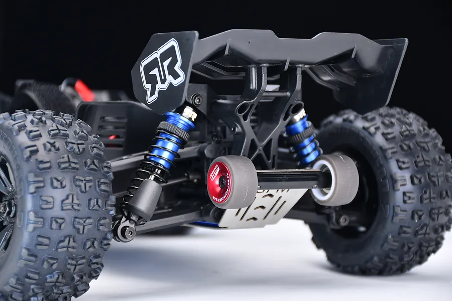 Rear view of an RC car chassis featuring a GPM Racing red aluminium wheelie bar installed below the rear wing and blue shock absorbers.