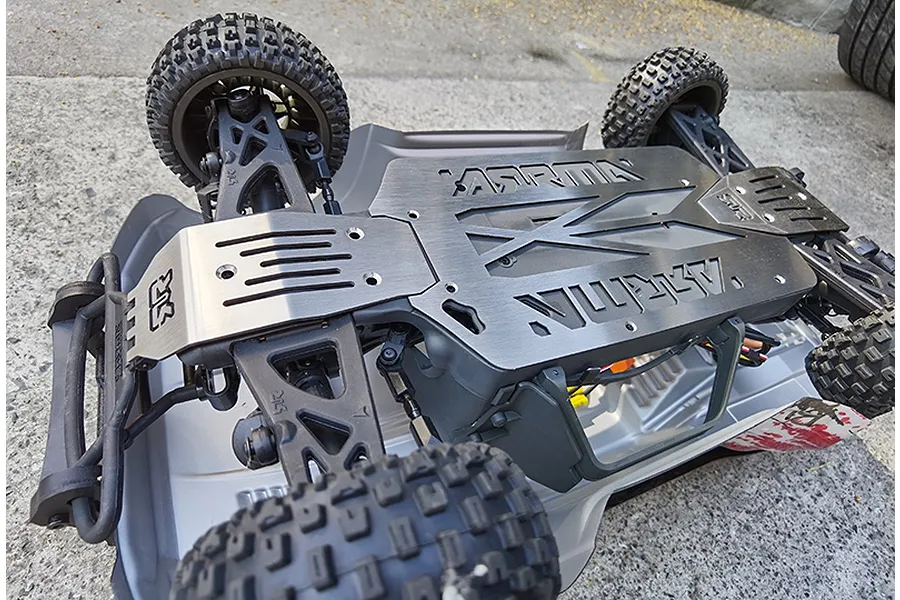 Underside view of an RC vehicle showing a GPM stainless steel chassis skid plate installed on an Arrma chassis.