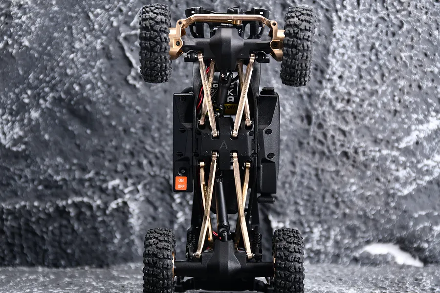 Bottom view of an RC crawler chassis featuring GPM brass weighted suspension linkages installed on a textured grey rock-like background.