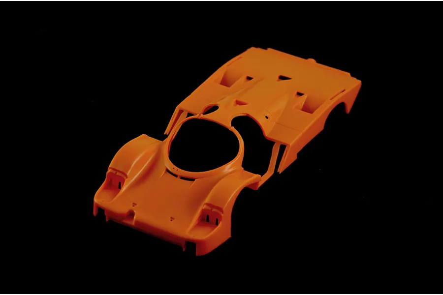 An unpainted orange plastic model car body shell for a Porsche 956, viewed from a high angle against a black background.
