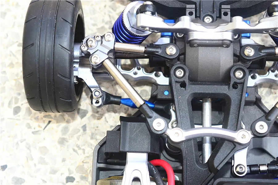 Close-up of an RC car front suspension assembly featuring GPM titanium tie rods and blue aluminium components installed on a chassis.