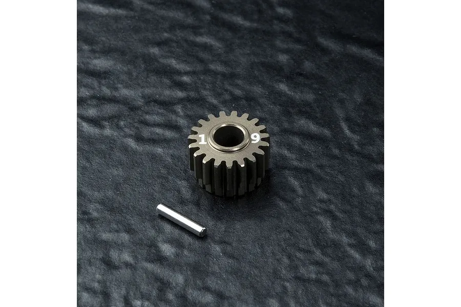 A 19-tooth metal transmission gear with the number 19 printed on its face, accompanied by a small metal pin, set against a dark textured background.