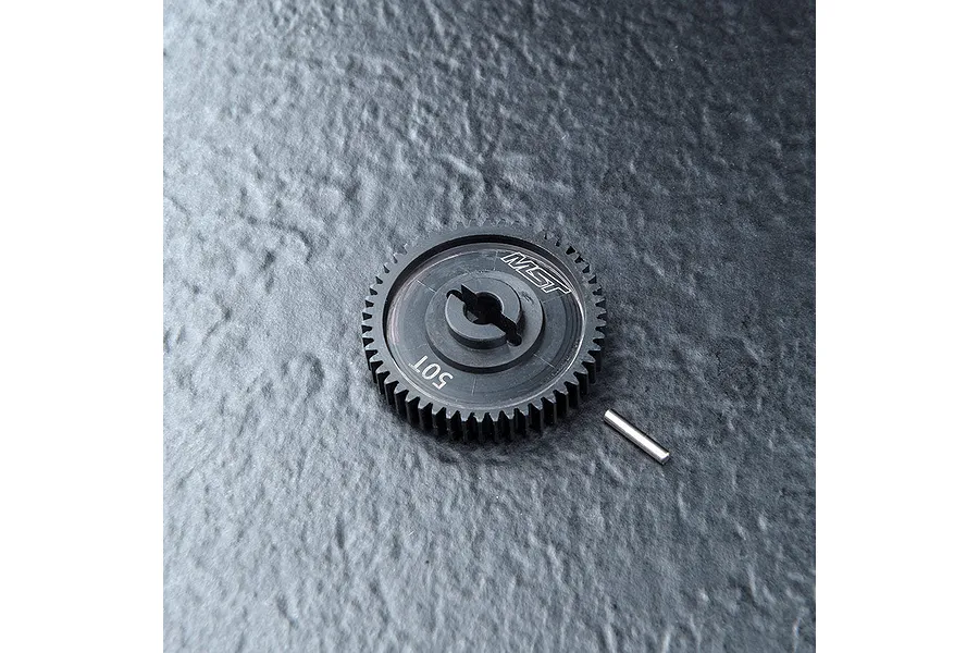 A black MST 50T spur gear with a small metal pin resting on a textured dark grey surface.