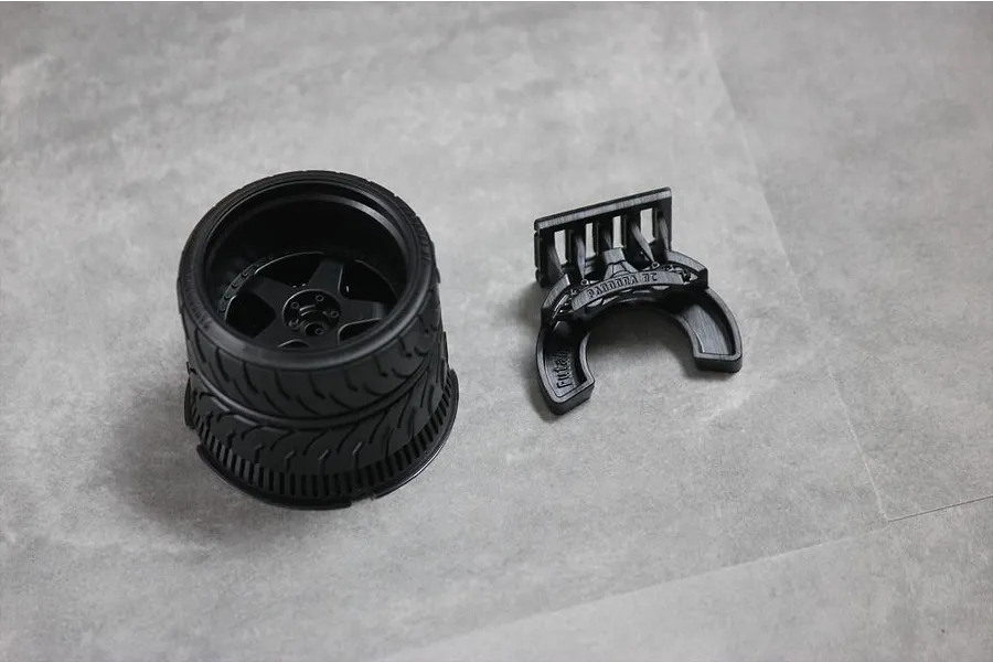 A black 3D-printed RC transmitter steering wheel assembly and a separate mounting bracket on a grey surface.