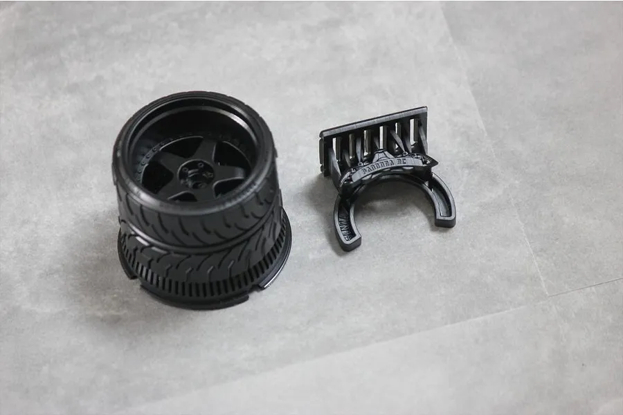 Black Pandora RC steering wheel accessory for Sanwa transmitters, featuring a detailed wheel rim and a separate mounting bracket on a grey surface.