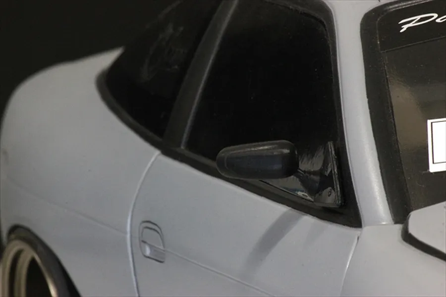 A close-up view of a black racing-style side mirror mounted on a light grey RC car body shell.