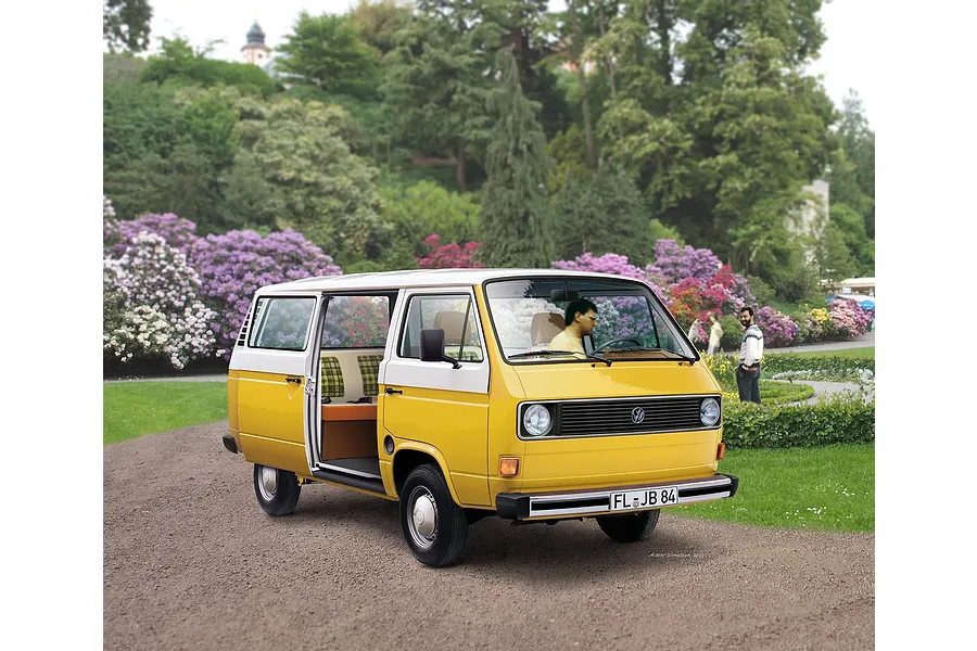 A yellow and white Volkswagen T3 Bus model car parked on a gravel path in a park with blooming flowers in the background.
