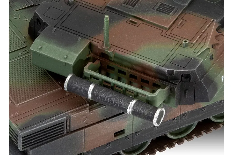 Close-up of a painted plastic model tank turret featuring camouflage paint, a storage rack, and a cylindrical component.
