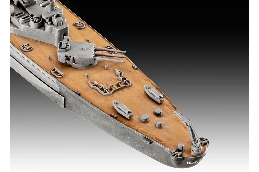 A close-up view of the bow section of a plastic model battleship, showing the wooden-textured deck, gun turrets, and small lifeboats.