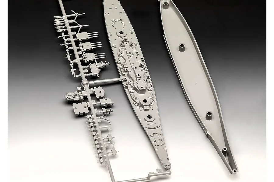 Unassembled grey plastic model kit parts for a battleship, including a sprue with turrets and the ship's deck and hull pieces.
