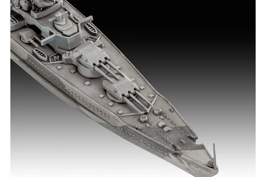 A high-angle close-up view of the bow section of a grey plastic model battleship, showing the deck, gun turrets, and anchor chains.
