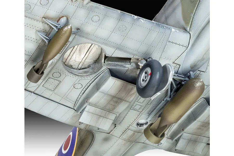 Close-up view of the underside of a 1/32 scale Supermarine Spitfire model, showing landing gear, panel lines, and underwing bomb payloads.