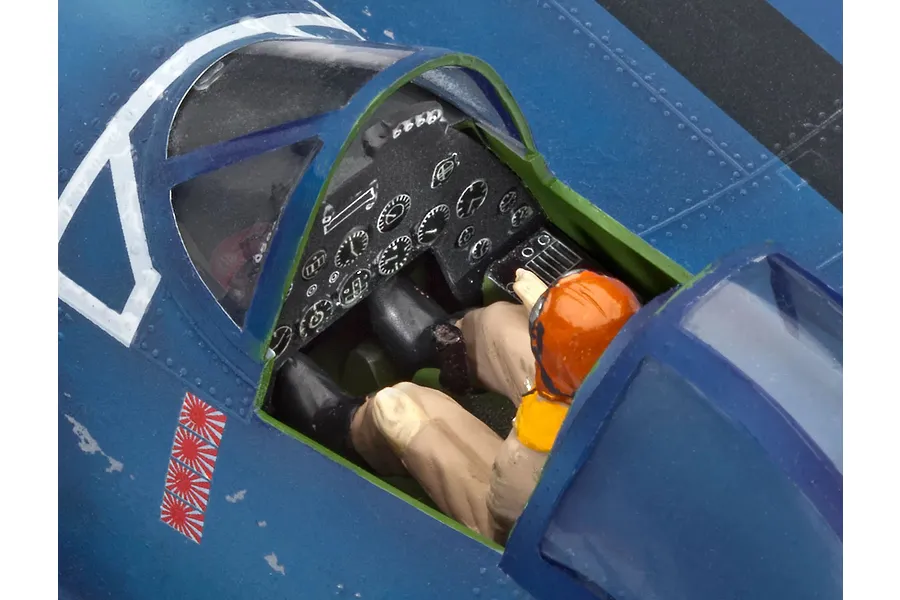 Close-up of a painted plastic model aircraft cockpit with a pilot figure seated inside, showing detailed instrument panel and blue fuselage.