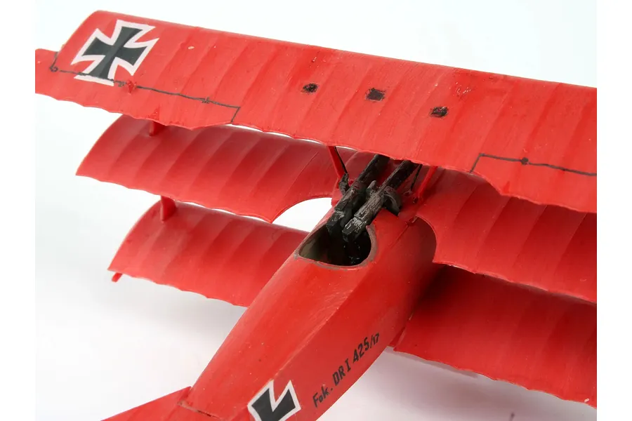A close-up, high-angle view of a red Fokker DR.1 triplane model aircraft showing the cockpit area, machine guns, and top wing markings.