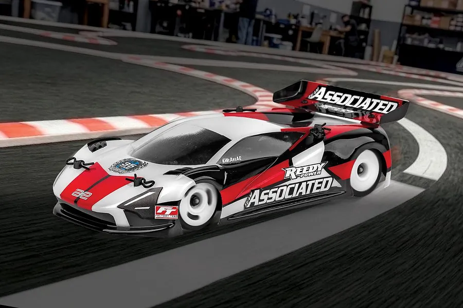 A Team Associated TC28 RC touring car driving on an indoor carpet race track with red and white curbing.