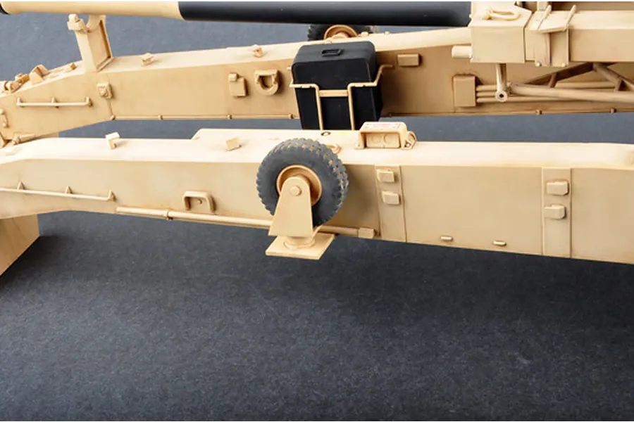 Close-up of the tan-colored chassis and support legs of a 1/16 scale M198 howitzer model kit, showing a small black storage box and a rubberized wheel.