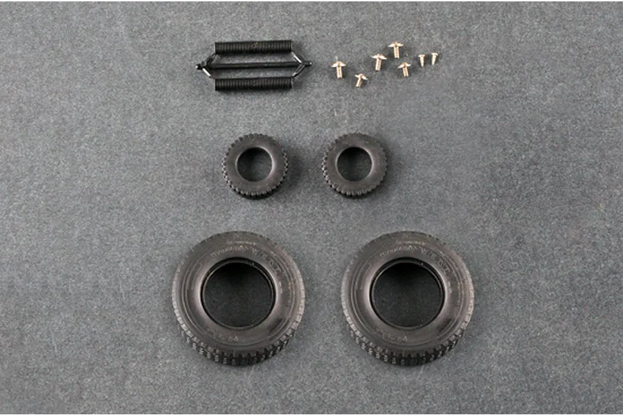 A top-down view of model kit parts including two large rubber tires, two small rubber tires, a black plastic spring assembly, and several small metal screws.