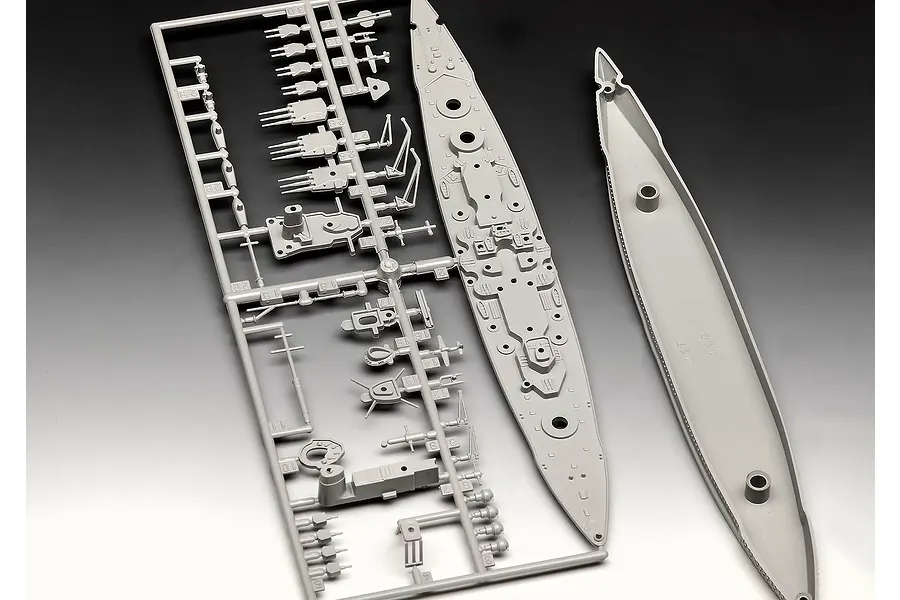 Grey plastic model kit parts for a battleship, including a sprue frame with turrets and the ship's deck and hull pieces.