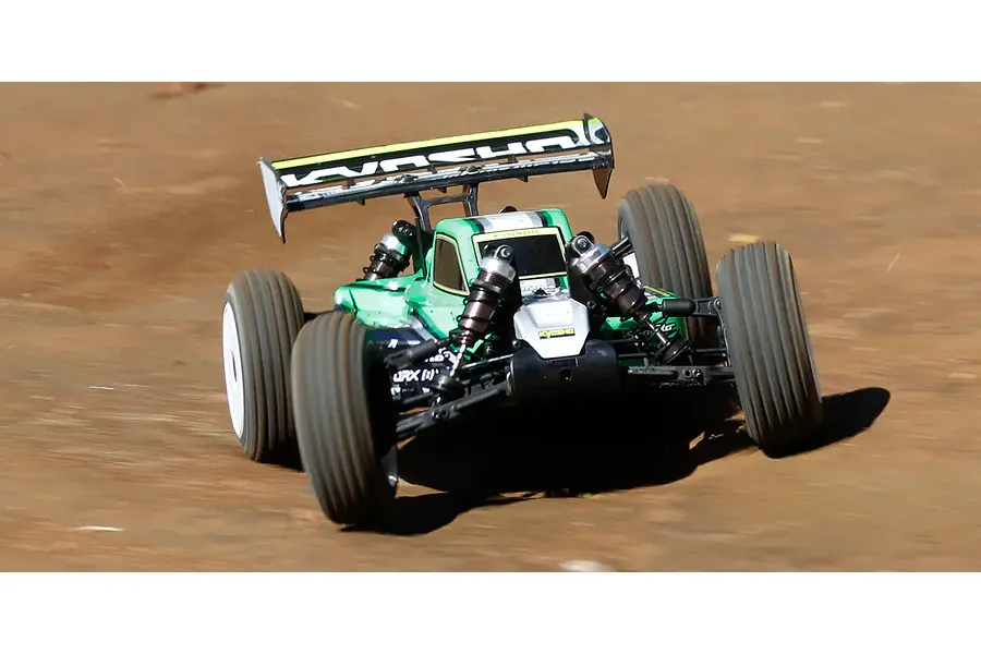 A green Kyosho Inferno MP10e RC buggy driving on a dirt track, showing its rear wing and suspension.