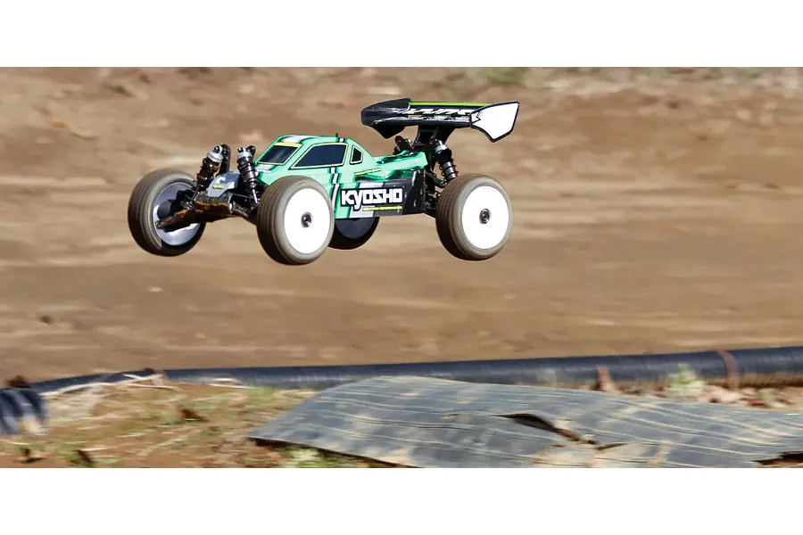 A green and black Kyosho Inferno MP10e RC buggy in mid-air during a jump on a dirt track.