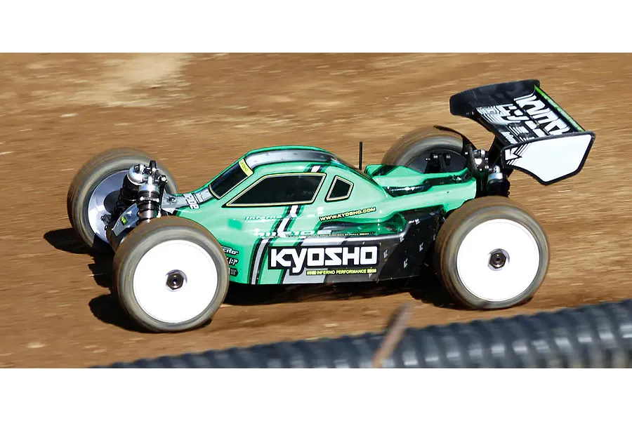 A green Kyosho Inferno MP10e 1/8 scale RC buggy driving on a dirt track, showing white wheels and black rear wing.