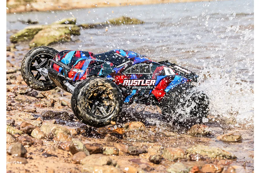 Traxxas Rustler Brushless RC stadium truck driving through shallow water, creating a large splash on a rocky shore.
