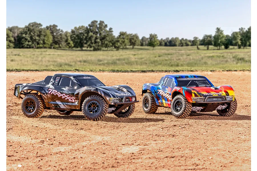 Two Traxxas Slash RC short course trucks, one black and one multi-coloured, parked on a dirt track outdoors.