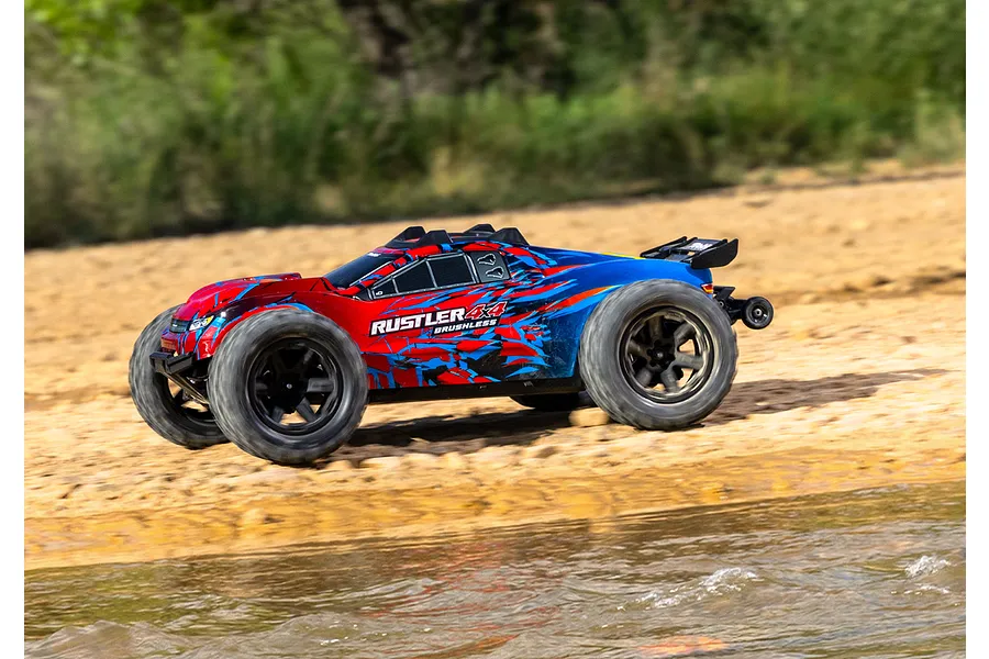 A Traxxas Rustler 4X4 VXL RC stadium truck with a red and blue graphic body driving along a sandy riverbank.