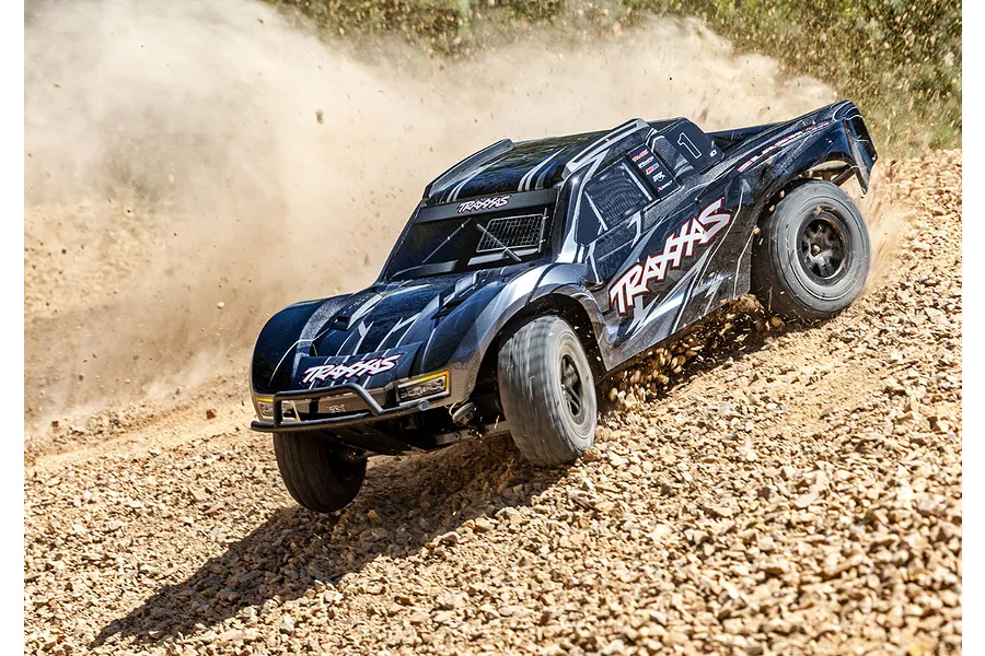 A black Traxxas Slash 4X4 RC short course truck driving fast on a rocky, dusty dirt track, kicking up a cloud of dust.
