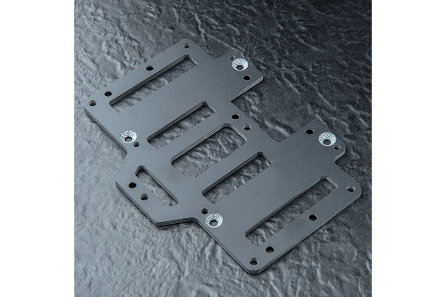 A black FRP battery mounting plate for an RC car, featuring multiple cutouts and countersunk mounting holes, set against a textured dark background.