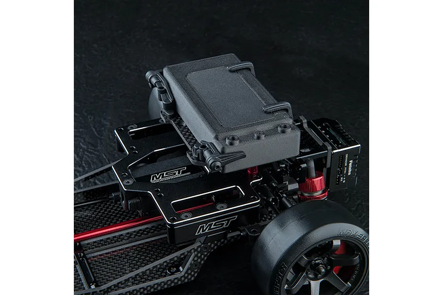 Close-up of an MST RC car chassis featuring a high-mounted battery tray with a black battery pack secured by clips.