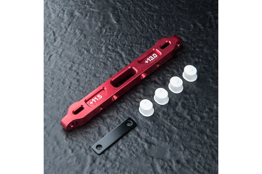 MST red anodized aluminium lower suspension arm mount with white plastic inserts and a black metal plate on a dark textured surface.