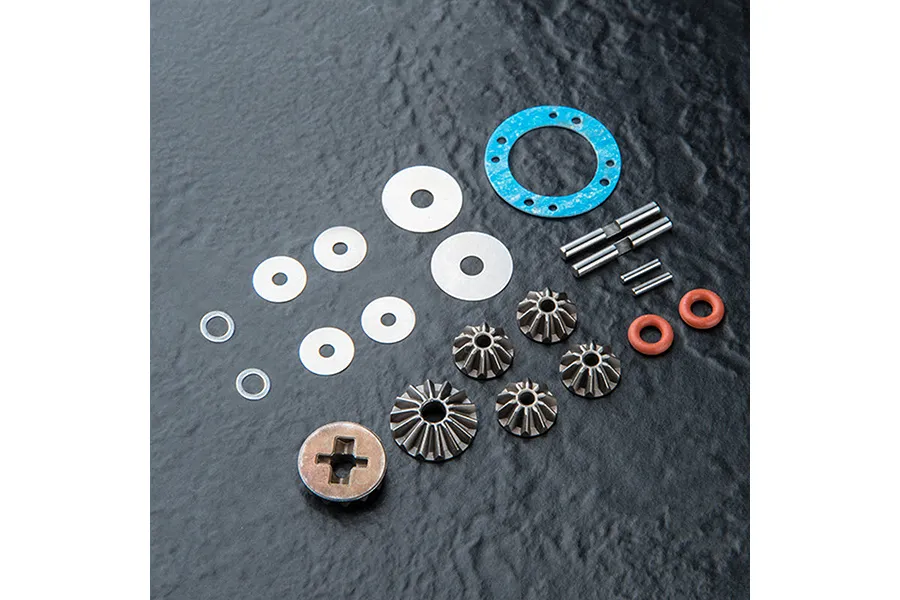 A set of MST limited slip differential parts including metal gears, washers, pins, O-rings, and a blue gasket on a dark textured surface.