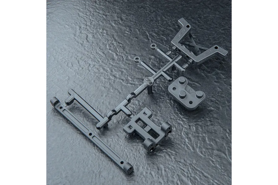 A black plastic parts tree containing various RC chassis components, including a shock tower and bumper mount, resting on a textured dark surface.