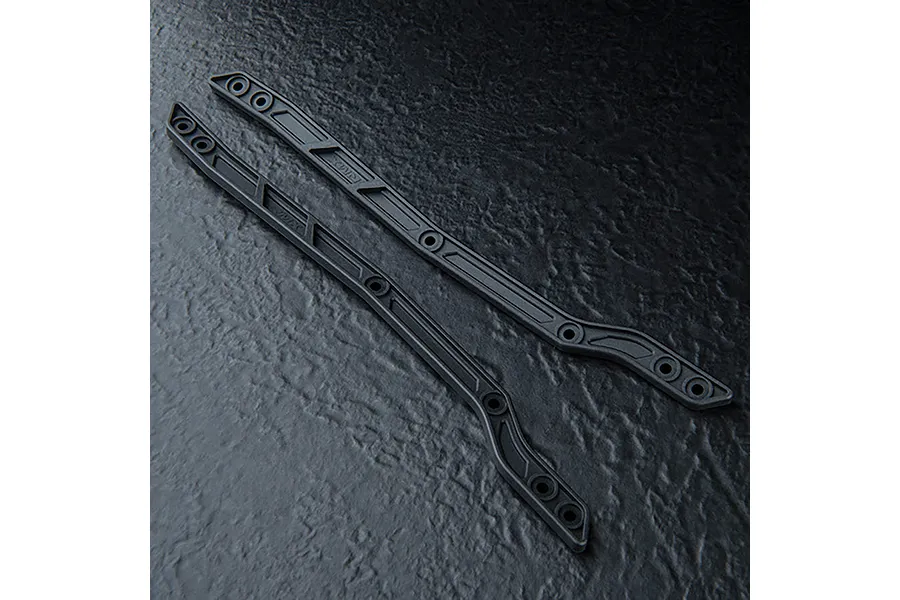 Two black MST RC chassis upper deck rails laid diagonally on a dark textured surface.
