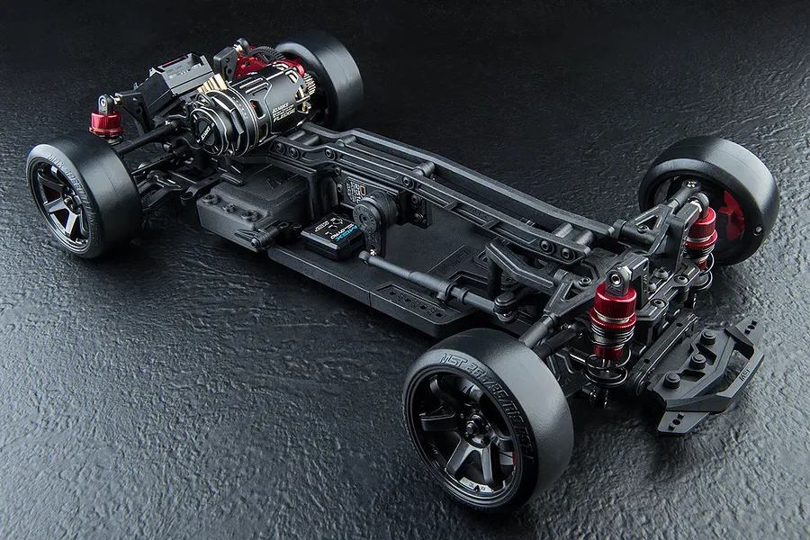 High-angle studio shot of an MST RMX 4 S Pro RC drift car chassis, showing the rear-mounted motor, suspension, and drift tires on a textured black surface.