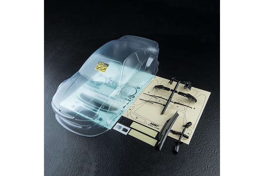 Clear, unpainted MST S15RB RC car body shell with included black plastic parts and decal sheet on a dark textured background.