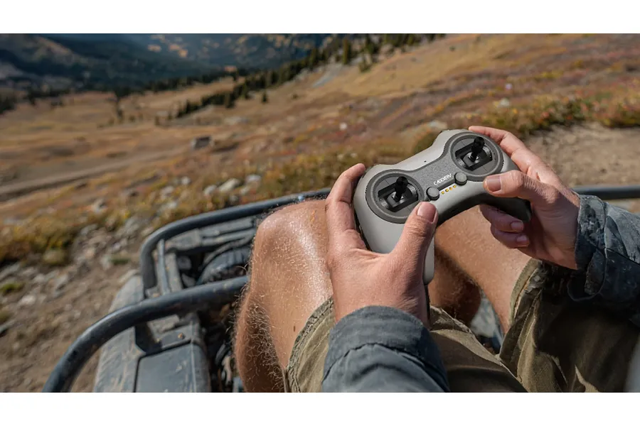 A person holding a grey CaddxFPV radio controller while sitting in an off-road vehicle with a mountain landscape in the background.