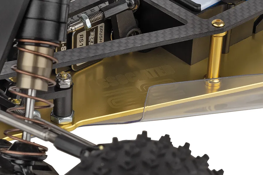 Close-up of the gold-anodized aluminum chassis of a Team Associated RC10 buggy, showing carbon fiber components and suspension details.
