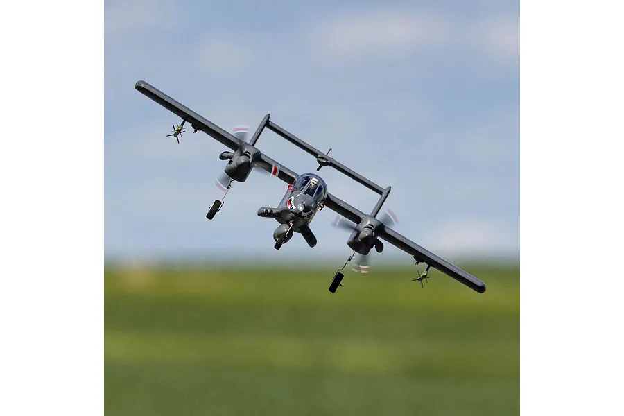 An E-Flite Micro OV-10 Bronco RC plane flying in the air with a blurred green field and blue sky background.