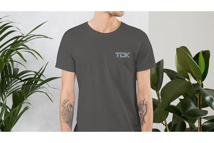 A person wearing a dark grey T-shirt with a TDK logo on the chest, standing between two indoor plants.
