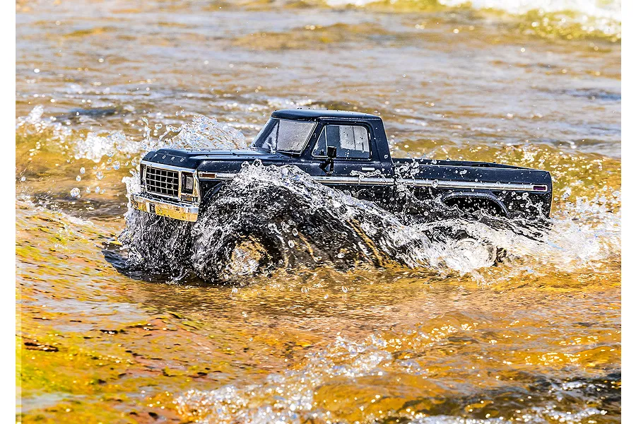 A black Traxxas TRX-4 1979 Ford F-150 RC truck driving through shallow, sunlit water, creating a large splash.