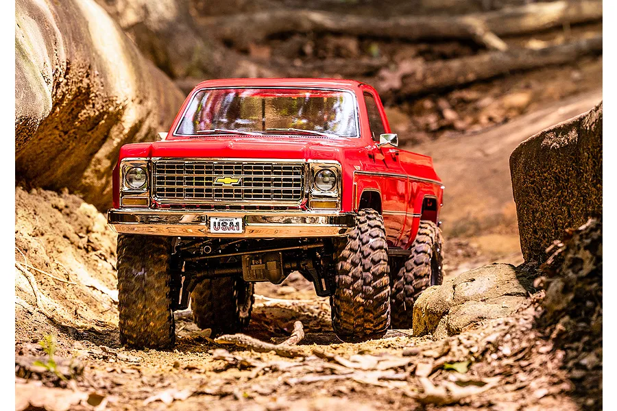 A red Traxxas TRX-4 1979 Chevrolet Cheyenne K10 RC crawler driving on a dirt trail between large rocks.
