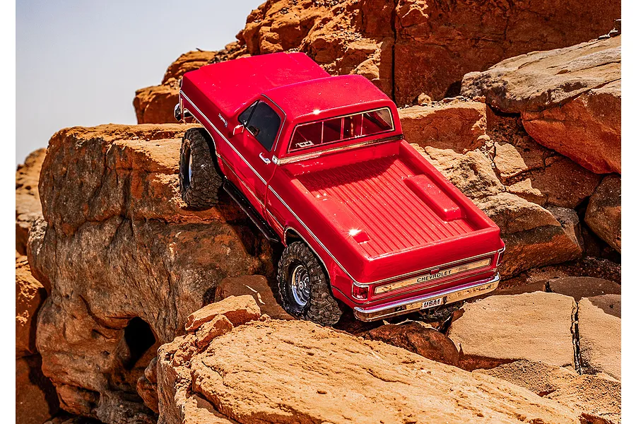 A red Traxxas TRX-4 1979 Chevrolet Cheyenne K10 RC rock crawler climbing over rugged orange rocks in an outdoor setting.