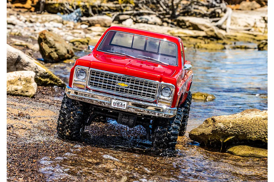 A red Traxxas TRX-4 1979 Chevrolet Cheyenne K10 RC rock crawler driving through shallow water over rocky terrain.