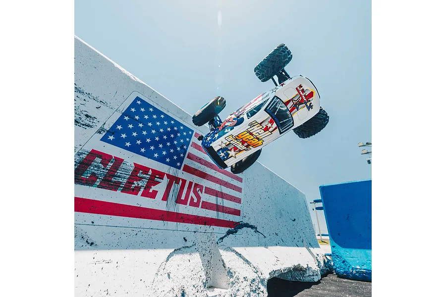 An ARRMA Freedom Flyer RC monster truck performing a high jump in front of a concrete barrier painted with an American flag and the word FLEETUS.
