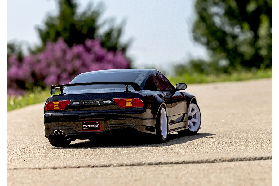 A black Traxxas Nissan 240SX RC car driving on a paved road with a blurred background of green trees and purple flowers.